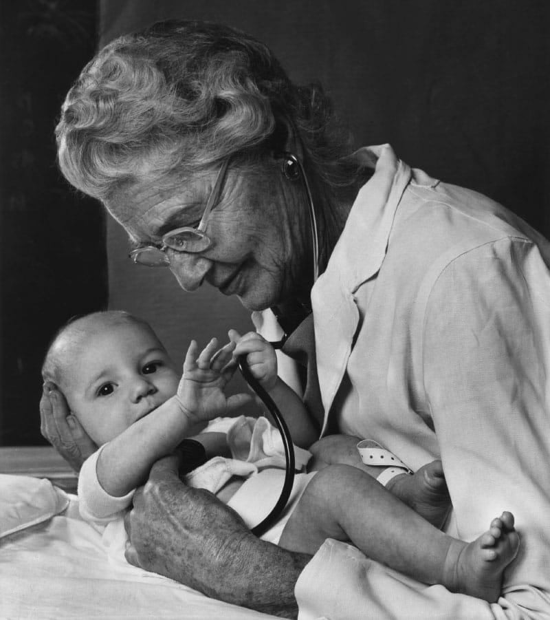 在这张1975年的照片中. 海伦·陶西格给婴儿戴听诊器. 在现实生活中，由于听力损失，陶西格很少使用这种乐器. (图片来源:Yousuf Karsh)